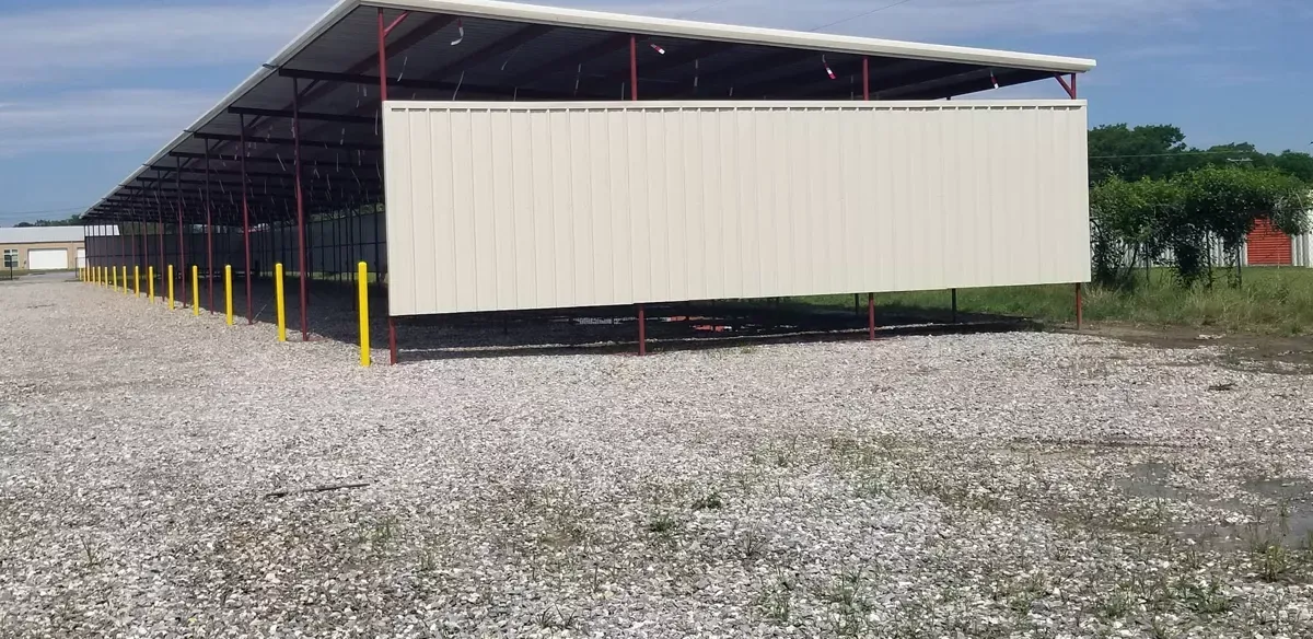 Long beige warehouse shed on gravel lot under a metal roof, with yellow posts and trees in the background