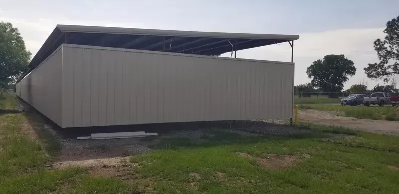 Long gray metal shed with a raised roof, sitting on a grassy lot under a cloudy sky