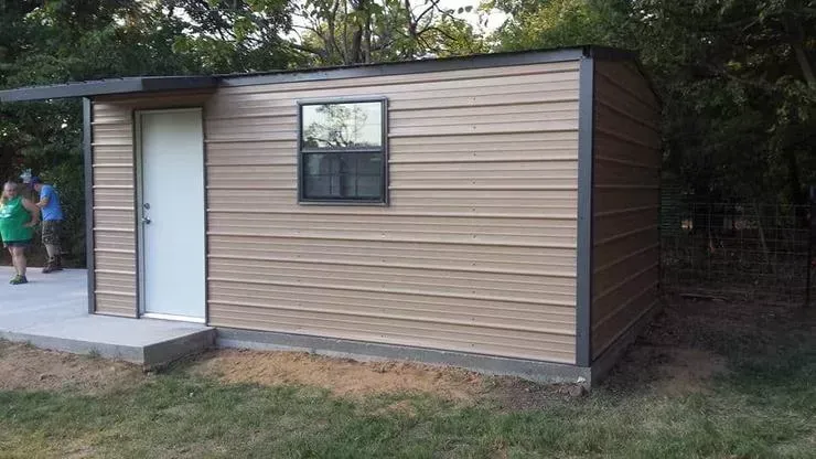 Small tan shed with white door and window in a grassy yard near trees