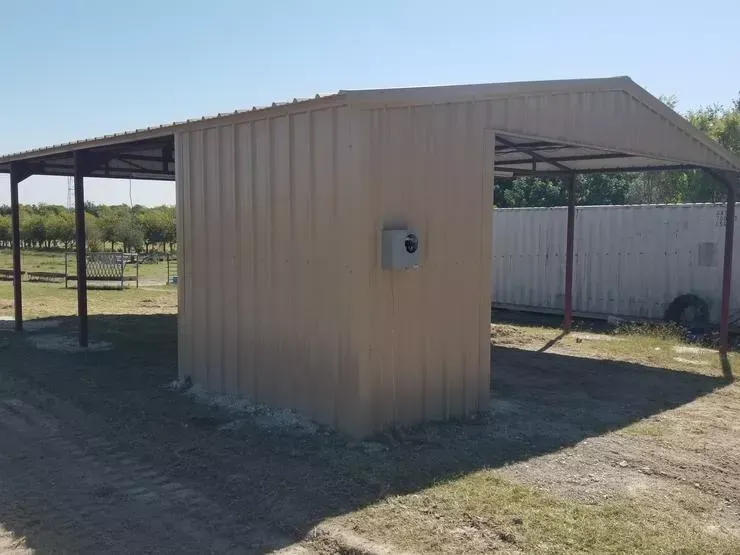 Tan metal shed with open sides on a grassy lot, with an electrical box on the wall.