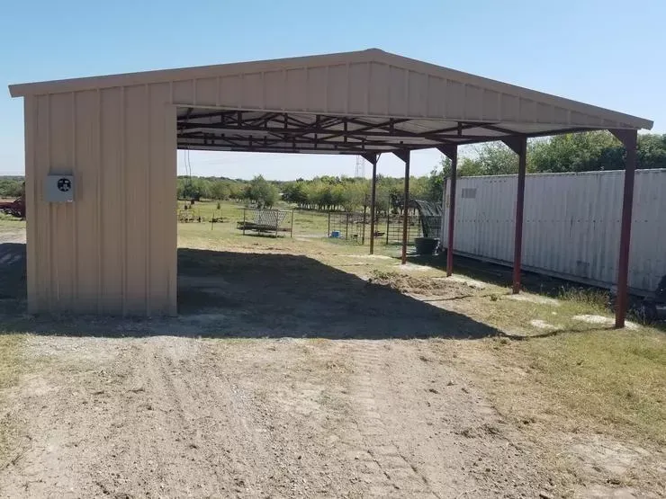 Open-sided metal shed beside a dirt road, with a beige wall and a fence in a rural yard.