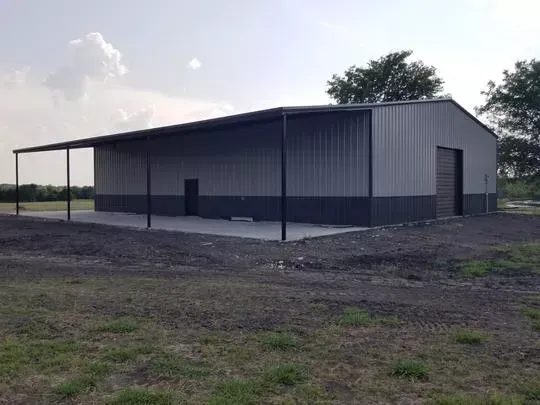 Gray metal barn-like building with a covered porch on a dirt lot under a cloudy sky