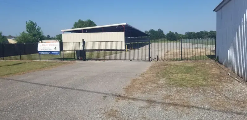 Closed gate at an industrial lot with a gravel driveway and white buildings under a blue sky