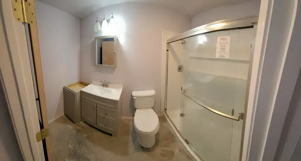Small bathroom with vanity, toilet, and glass shower enclosure, lit by a wall mirror.