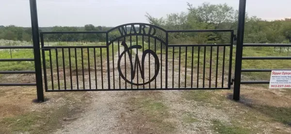 Black metal ranch gate with circular monogram, opening to a field under cloudy sky.