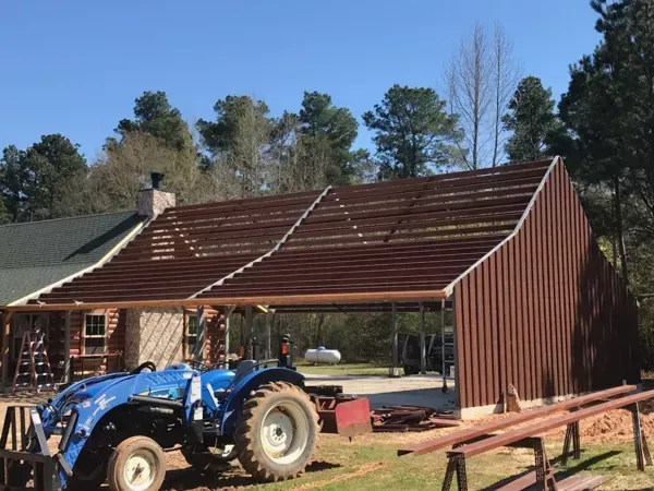 Blue tractor beside a red metal barn under construction in a wooded yard
