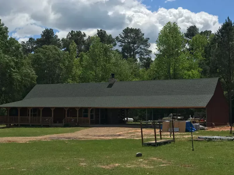 Single-story red cabin with dark roof on a grassy lot, trees and cloudy sky behind it