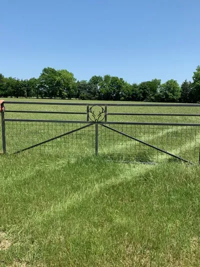 Black metal farm gate in a green grassy field with trees and blue sky behind.