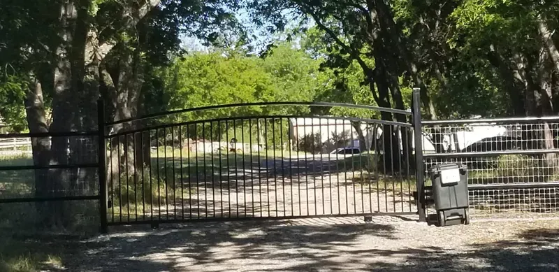 Black metal gate across a driveway under trees, with a small wheeled bin on the right