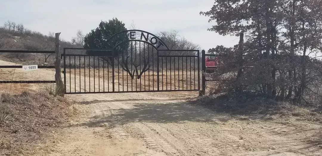 Closed black metal gate on a dirt driveway in a dry, rural area with trees and sparse brush.