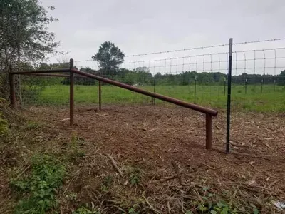 Rustic metal farm gate with slanted top bar beside a fenced green field under cloudy sky