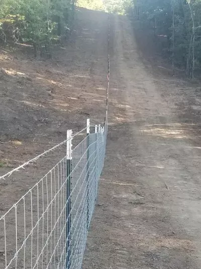 Dirt path or driveway beside a wire fence on a wooded hillside.