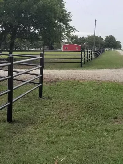 Black fenced paddock with grassy path and red barn in the distance under cloudy sky