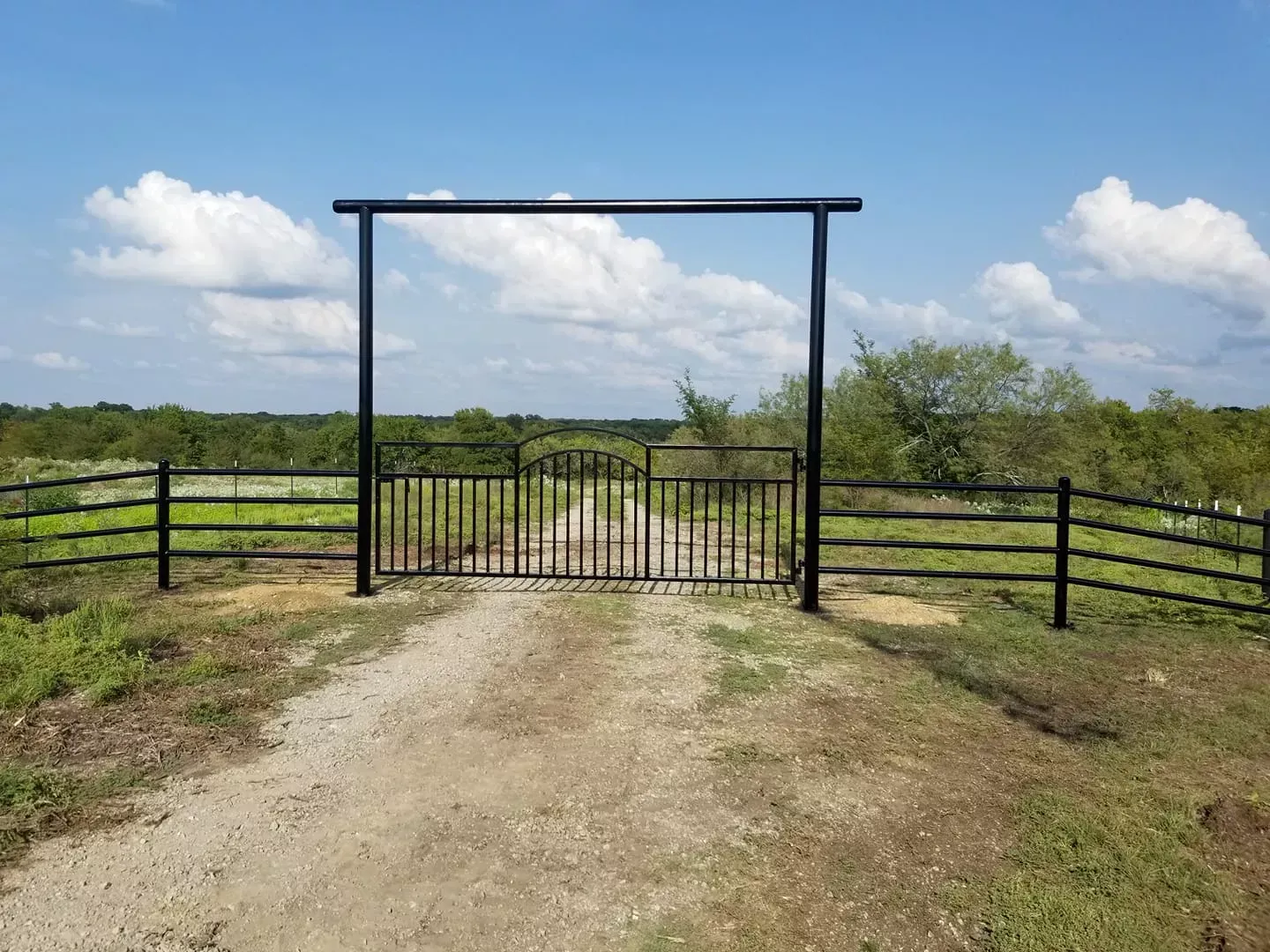 Gravel path leading to a closed black metal gate between fences in a green rural field under a cloudy sky