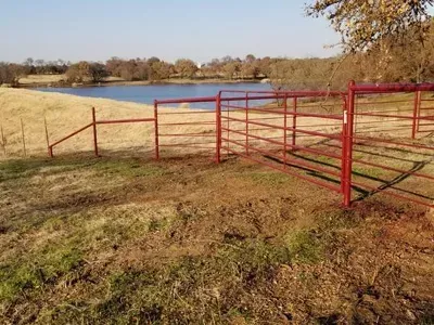 Red metal farm gate beside a pond, with dry grass and trees in the background.