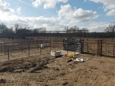 Empty livestock pen with metal fencing, feeding equipment, and muddy ground under a cloudy sky