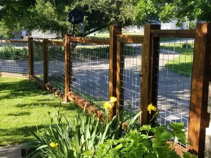 Wooden and wire garden fence beside a sunny grassy path with yellow flowers and trees
