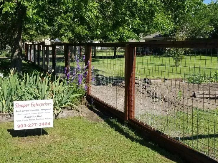 Wire fence enclosing a grassy yard with flowers, trees, and a small sign near the entrance.