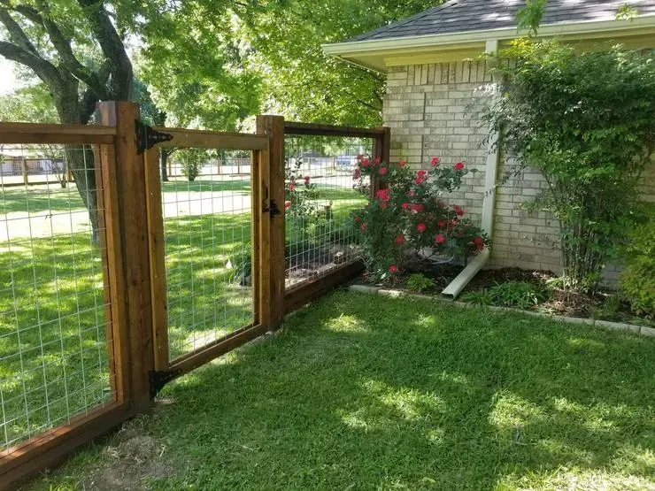Wooden gate and wire fence beside a stone house, with green lawn and pink flowers in a sunny yard