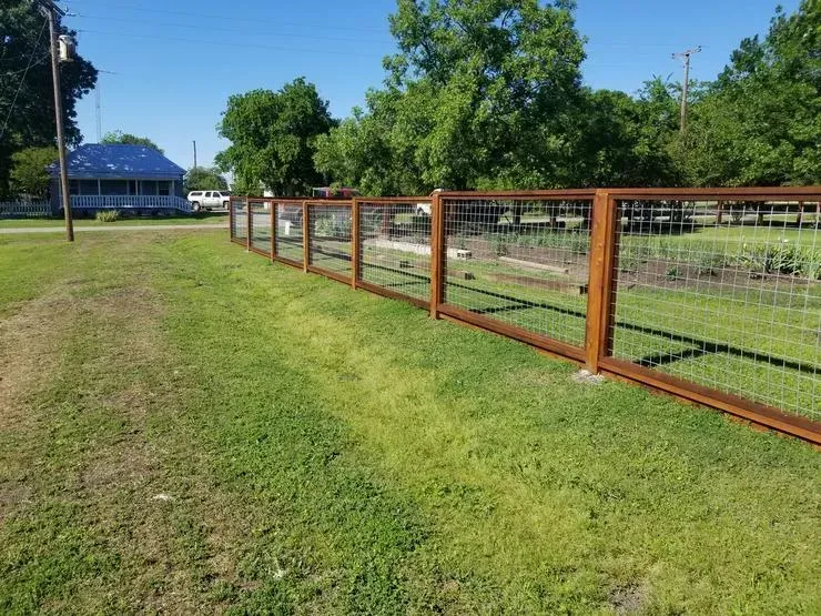 Rustic fenced pasture beside a grassy path, with trees and a blue-roofed barn in the distance.