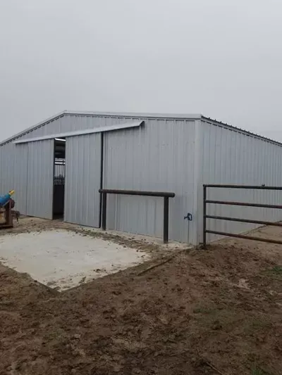 Gray metal barn with a large sliding door and fenced dirt yard under an overcast sky