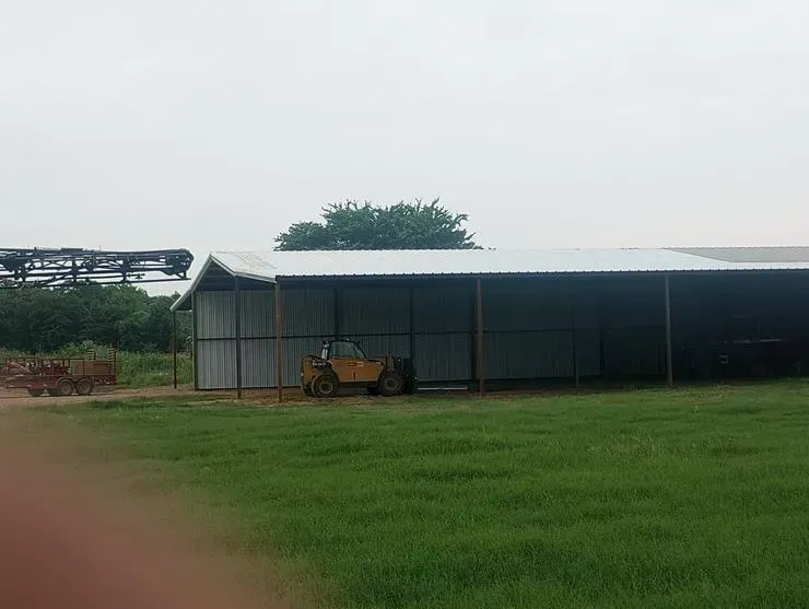 Small shed in a grassy field with a yellow tractor parked underneath a corrugated metal roof.