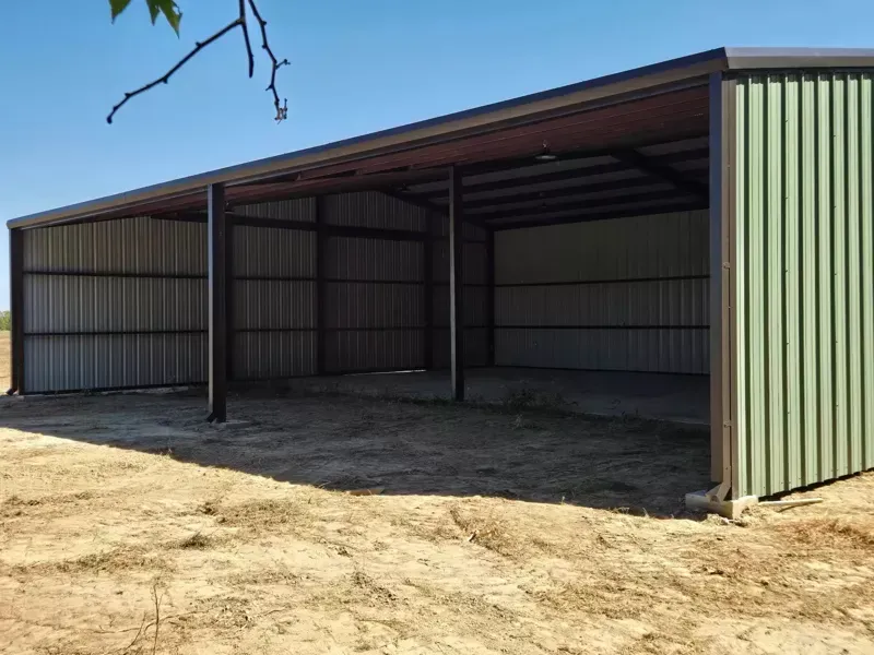 Open-sided green metal barn or shed with a dirt floor under a clear blue sky