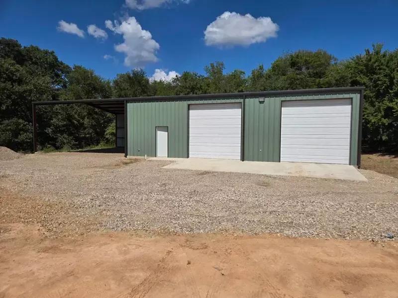 Green metal workshop with two white roll-up doors and a small side door on a gravel lot under a blue sky