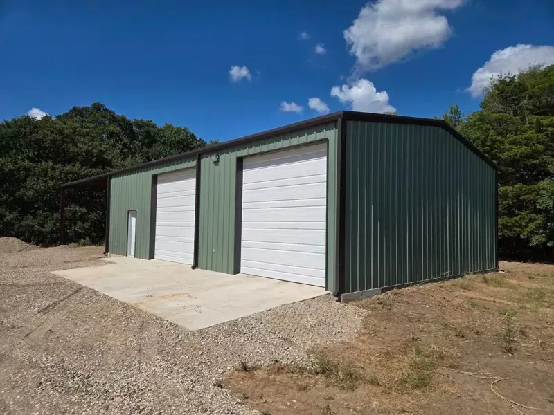 Green metal garage building with three white roll-up doors on a concrete pad under a blue sky