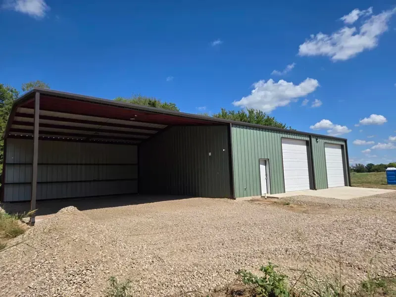 Green metal barn with open lean-to, gravel lot, and white garage doors under a blue sky