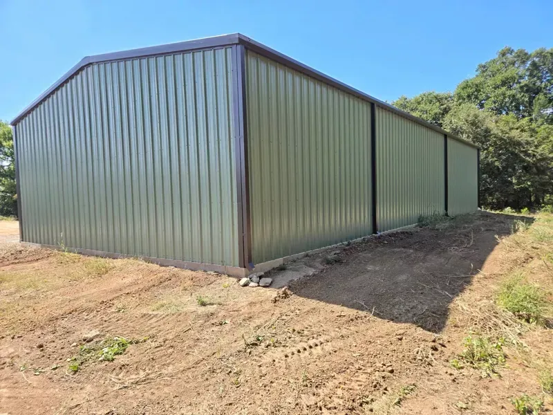 Green metal barn or shed on a dirt lot beside trees, viewed from the corner.