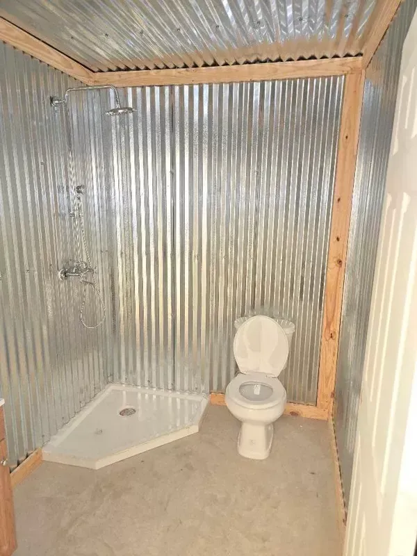 Small corrugated metal bathroom with a toilet and shower base, unfinished interior.