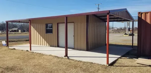 Small beige shed with red posts and a covered concrete porch in a grassy field
