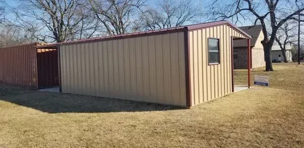 Tan metal shed with beige trim on a grassy lot beside a wooden fence and bare trees