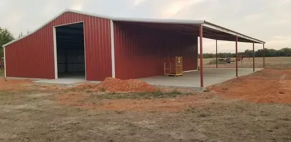 Red metal farm shed with open front, on a dirt lot under a cloudy sky.