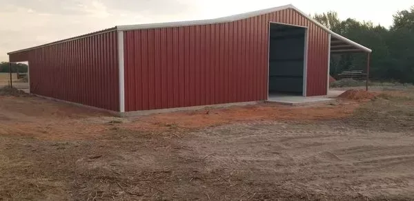 Red metal barn with large open doorway on a dirt lot, under a cloudy sky.
