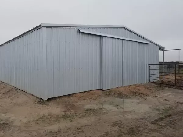 Gray metal barn or storage shed with large sliding doors on a dirt lot under an overcast sky