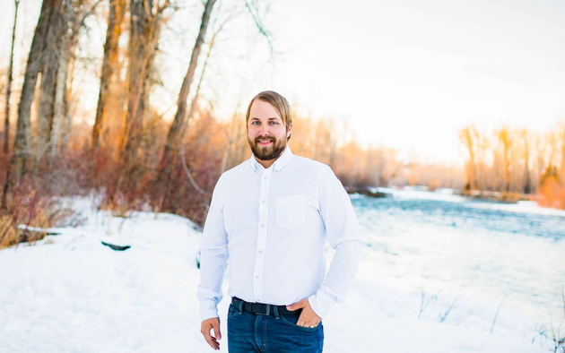 A bearded man in a white button-down shirt and blue jeans stands outdoors in a snowy landscape near a river at sunset.
