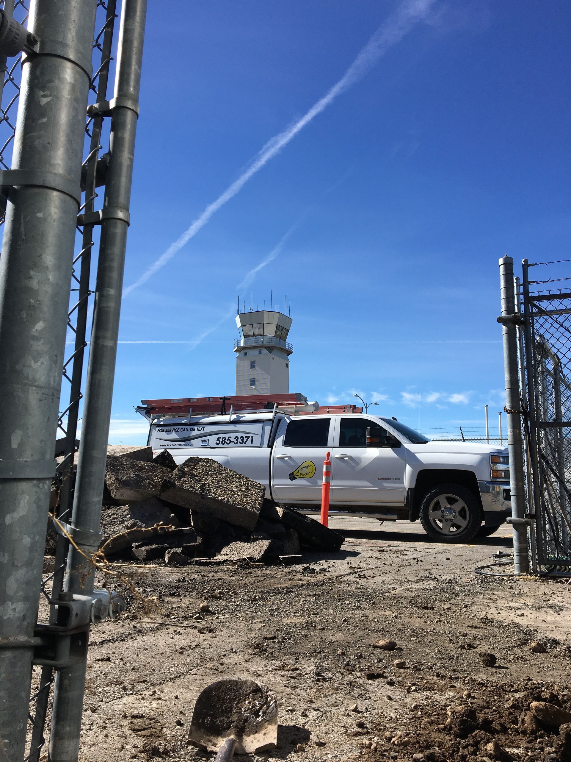 A white service truck parked on a construction site near an airport control tower under a clear blue sky.