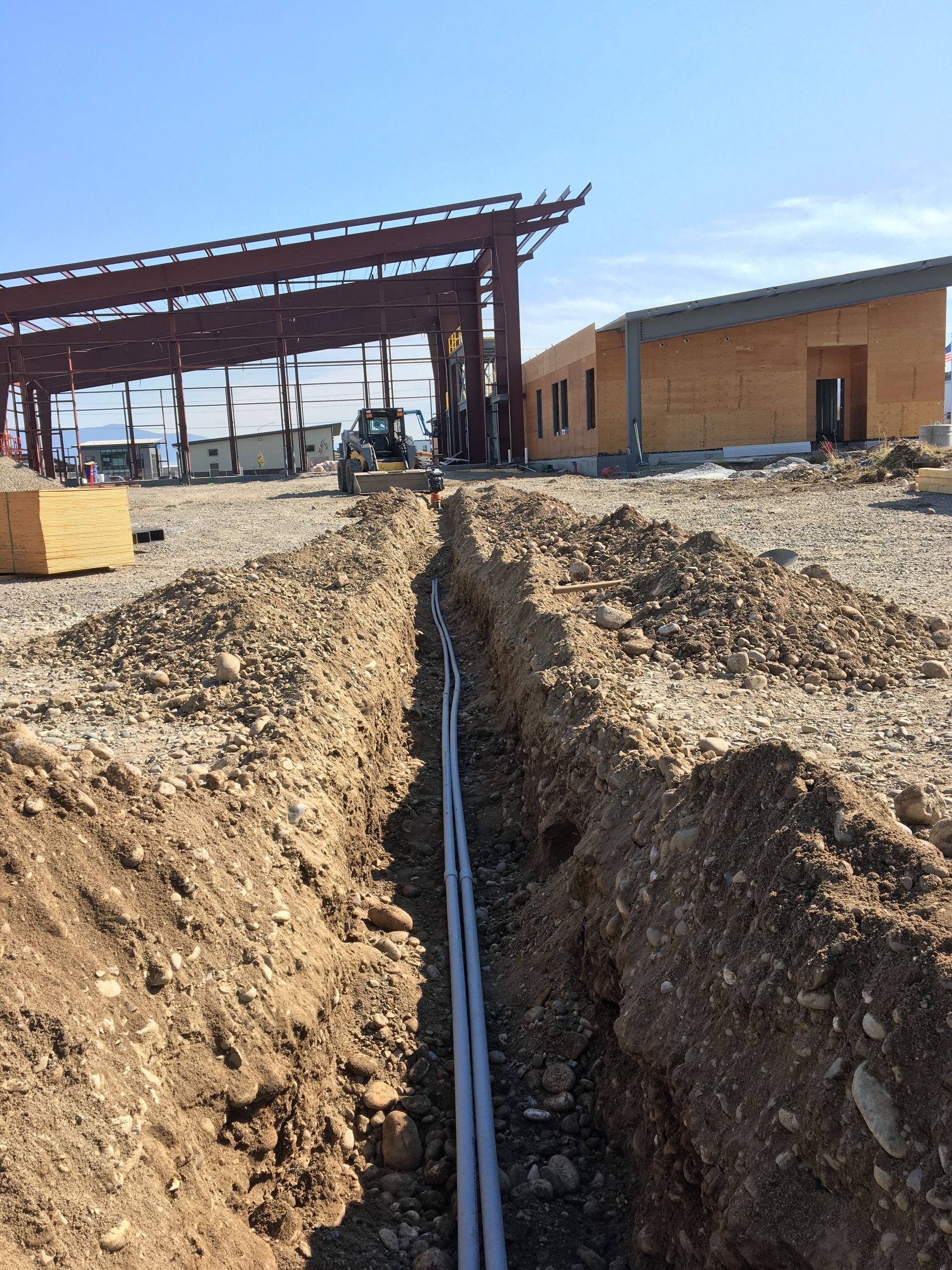 A narrow trench with gray conduit pipes running through a construction site towards two buildings under a clear sky.