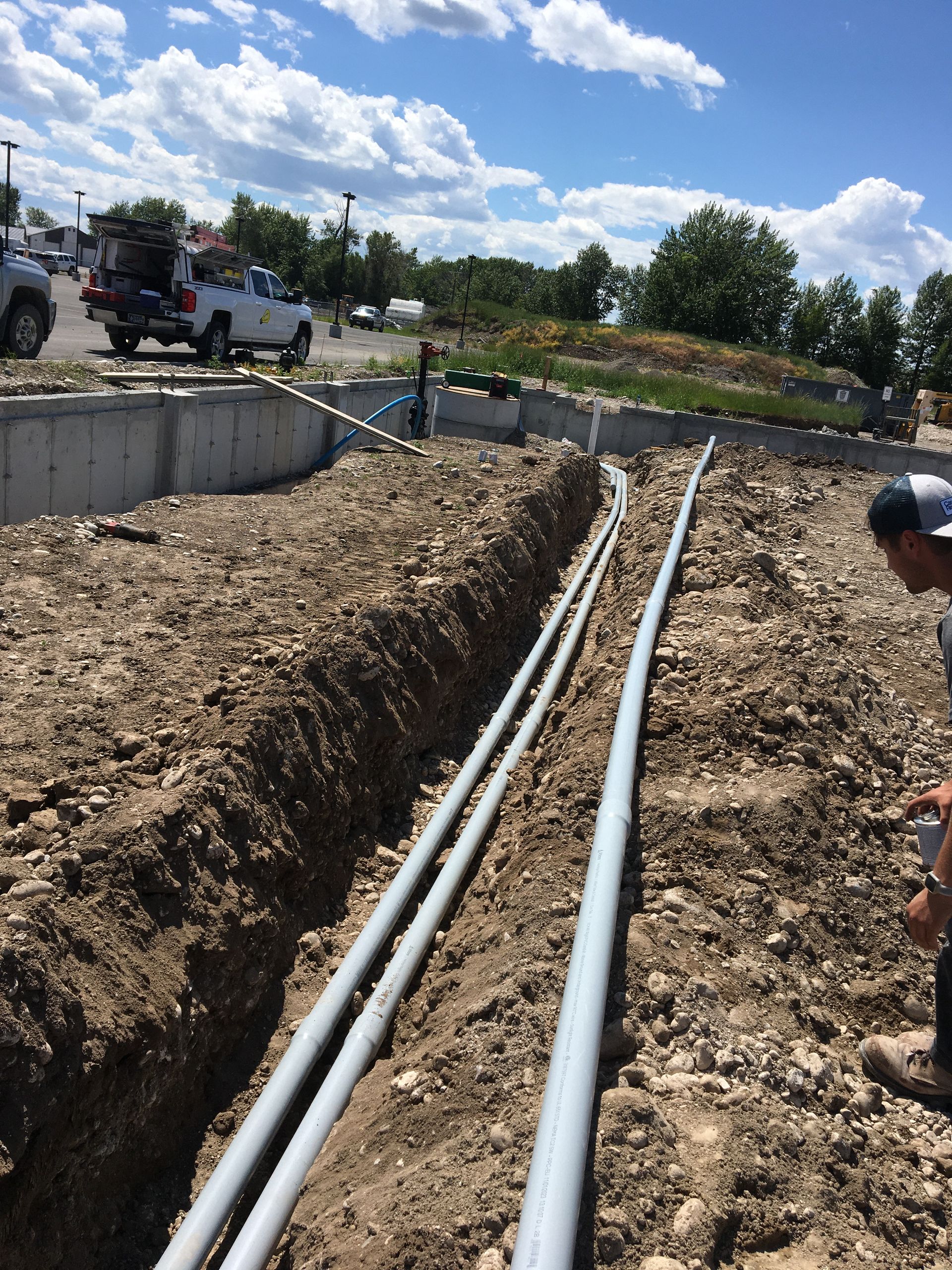 A person observes gray electrical conduits laid in a dirt trench at an outdoor construction site.
