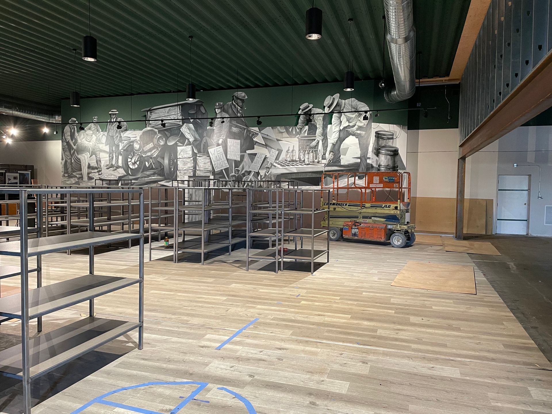 Empty metal shelving stands on a wood-look floor in a room featuring a large, monochrome mural of people on a green wall.