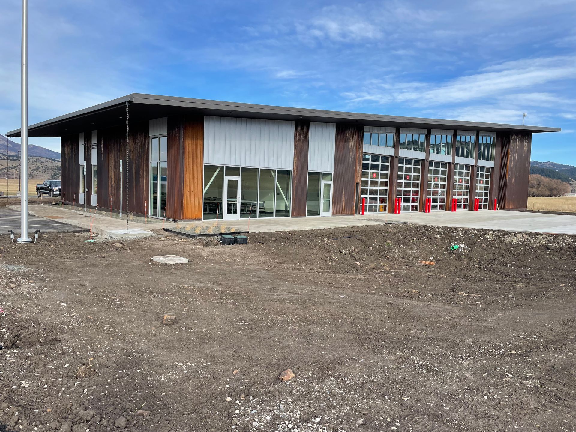 A modern fire station with rusted metal siding, large glass bay doors, and a flat roof against a bright blue sky.