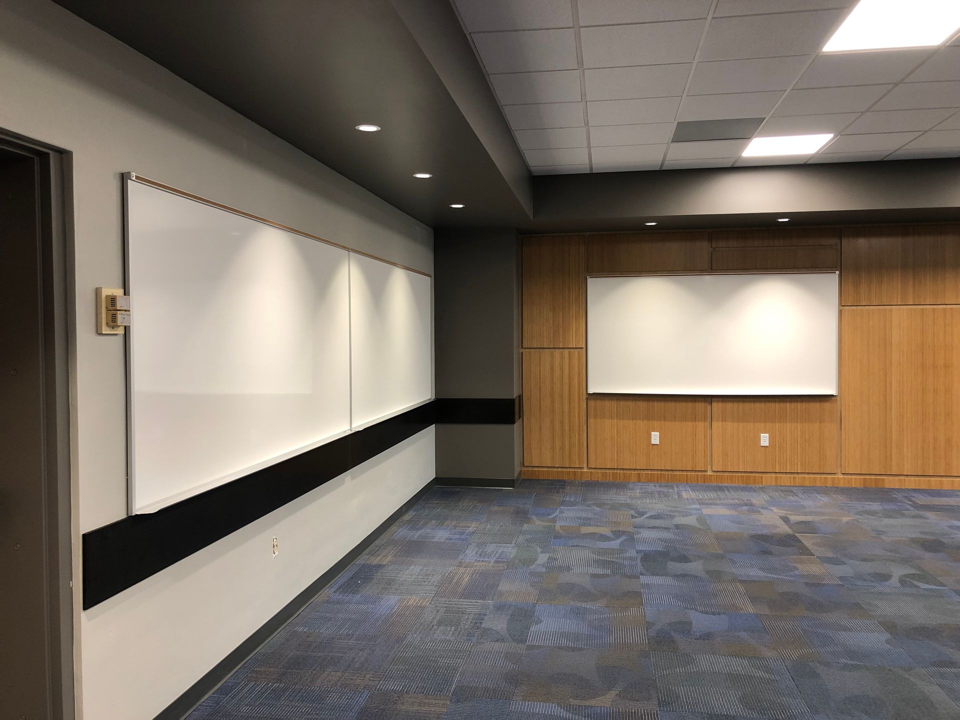 An empty modern office or conference room with whiteboards, wood-paneled walls, and blue patterned carpet.