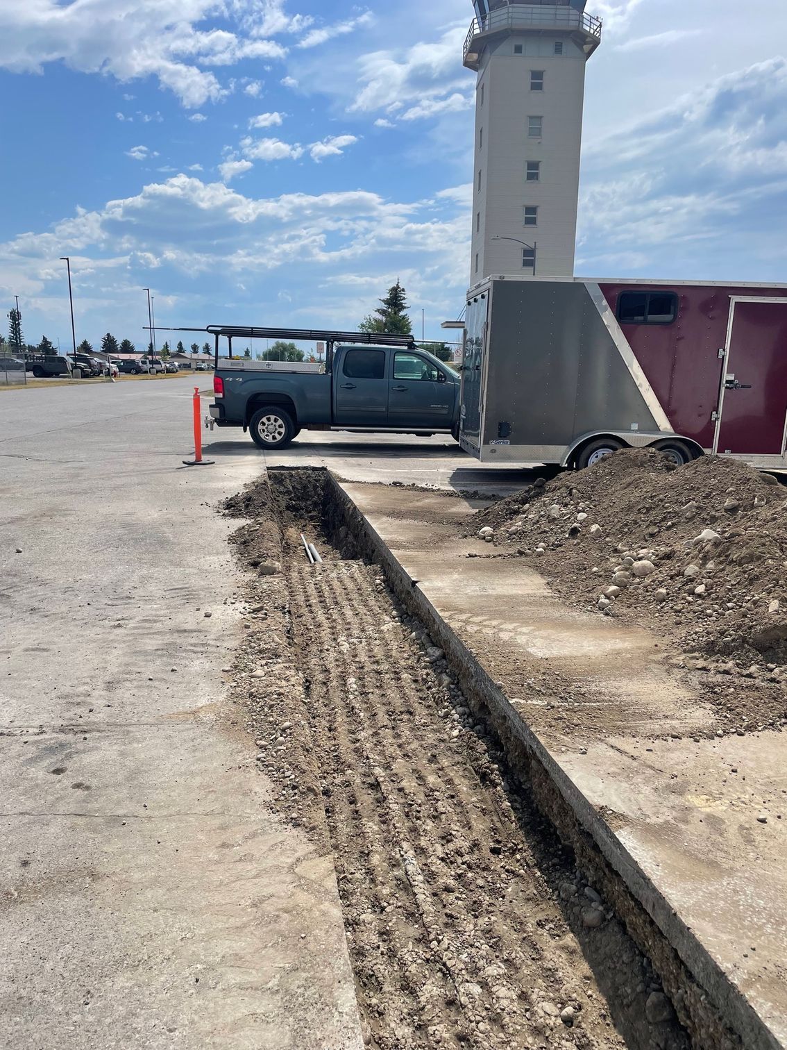 A trench cut into a paved lot next to a pile of dirt, a gray truck with a trailer, and a tall tower in the distance.