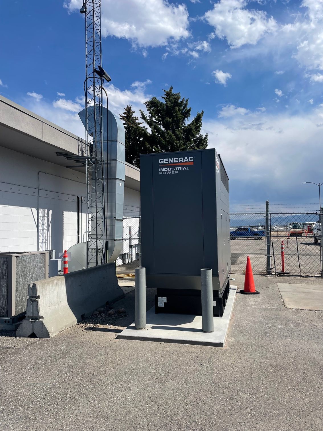 A grey Generac industrial backup generator sits on a concrete pad next to a building, with a vertical exhaust duct nearby.