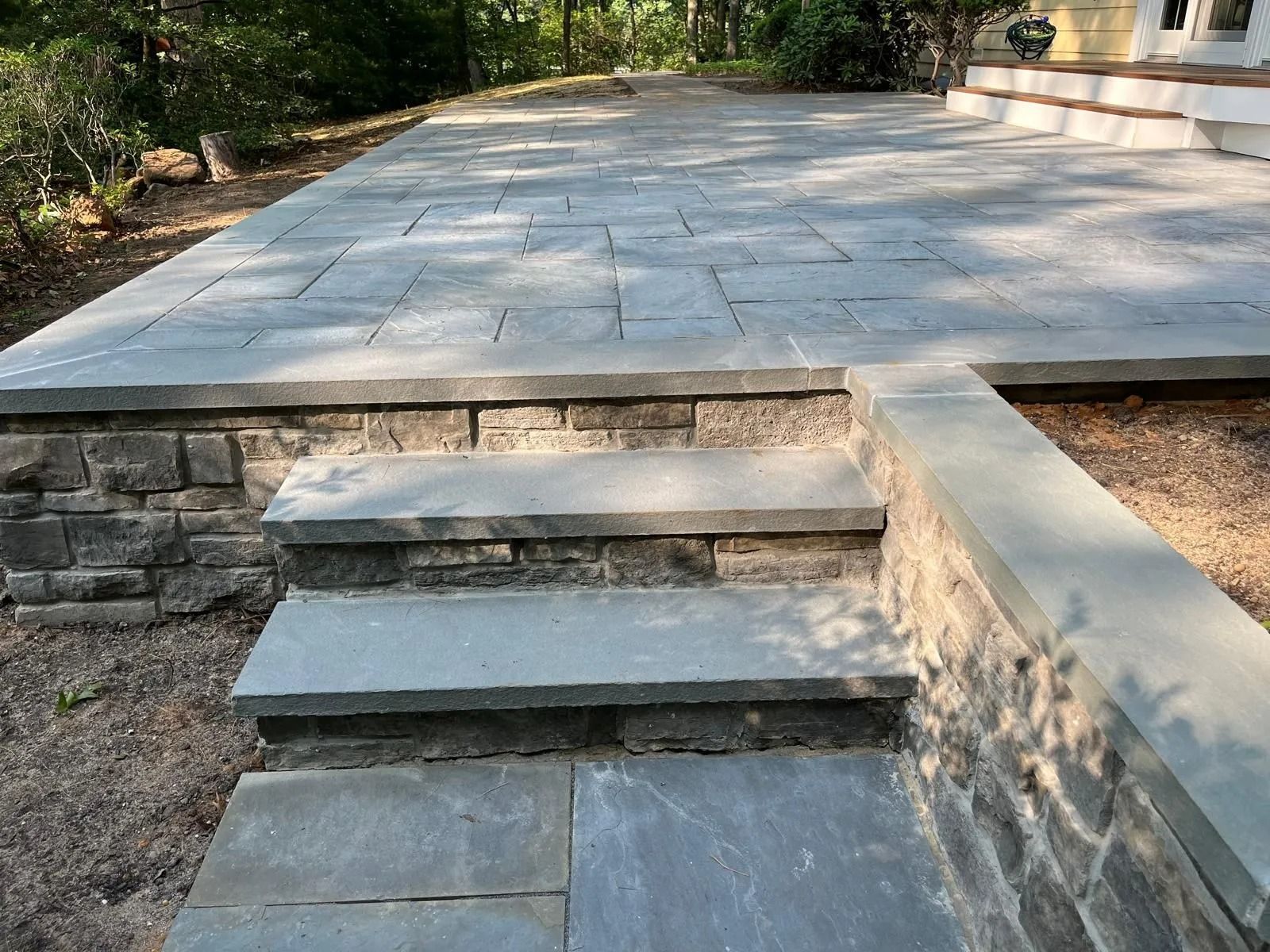 A gray stone patio features two stone steps leading up to an elevated, flagstone-paved seating area surrounded by trees.