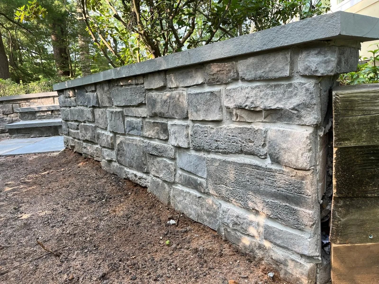 A low, gray stone retaining wall with a smooth capstone, situated in a garden setting with dirt in the foreground.