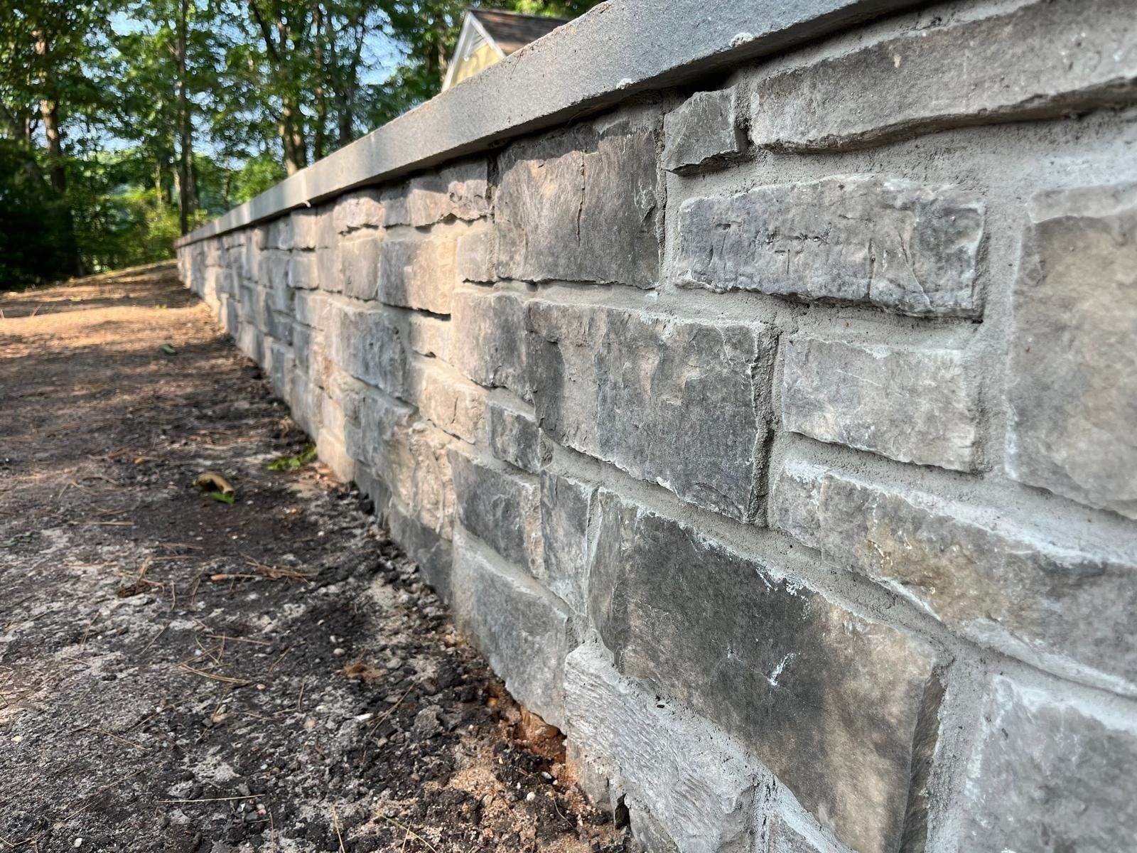 A stone retaining wall with a flat capstone topper runs alongside a dirt path near trees.