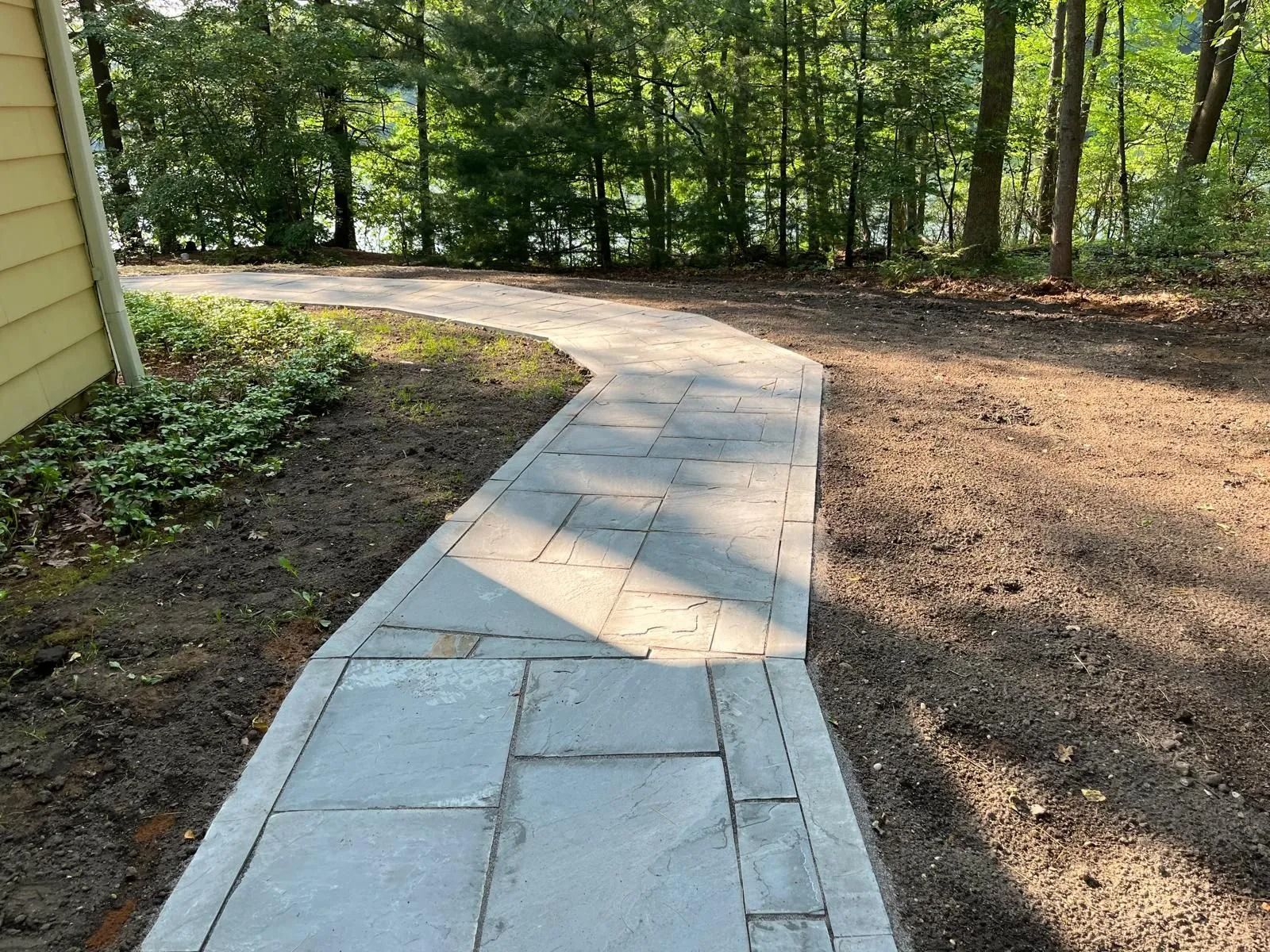 A gray stone walkway curves through a wooded yard toward a yellow house wall.